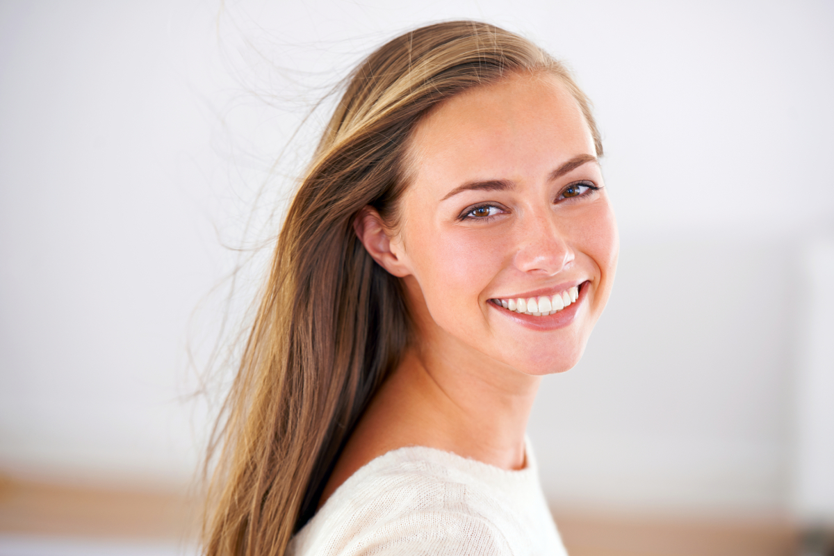 A smiling woman with long, flowing hair, wearing a light sweater, poses against a soft, neutral background.